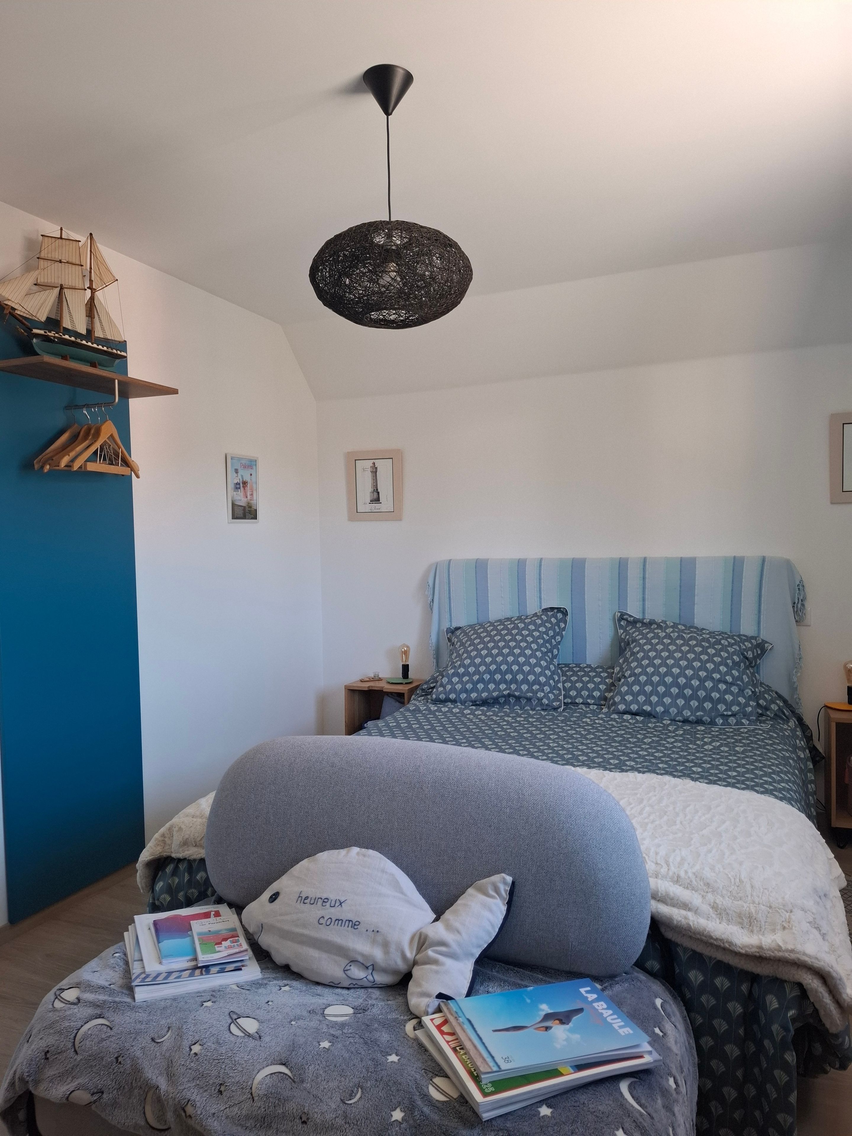 Quarto com cama, travesseiros, luminária pendente, prateleira com modelo de navio e parede com quadros.