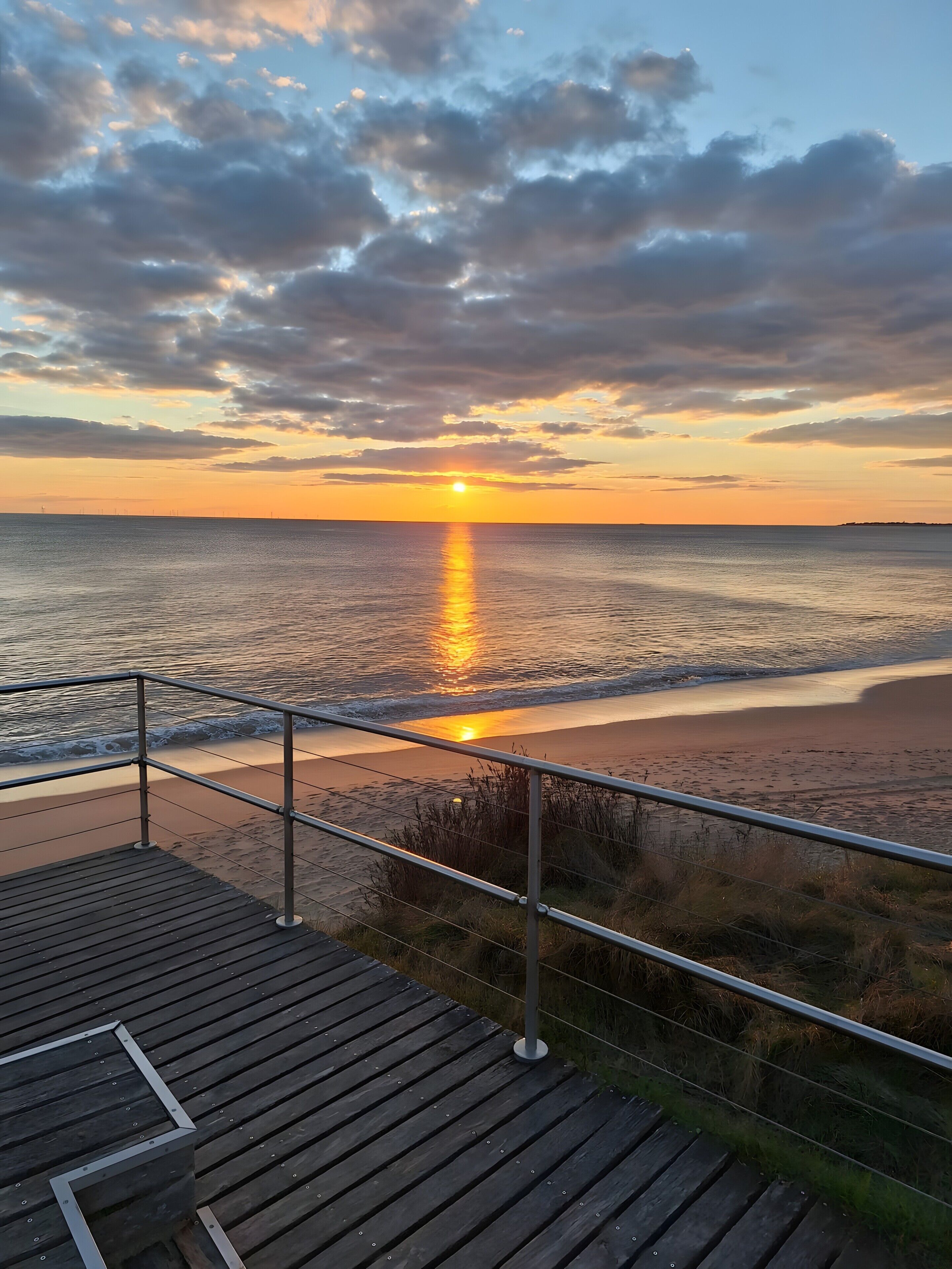Une terrasse en bois avec une rambarde, donnant sur une plage au coucher du soleil.