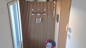 A compact room with a striped wall, a small shelf with decorative items, a door with a handle, a pair of slippers, and a small trash bin. - Paloma by the Sea Guest Room with Shared Terrace, Garden, and Wi-Fi (Pornichet)