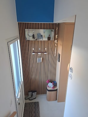 Una camera compatta con una parete a righe, un piccolo ripiano con oggetti decorativi, una porta con maniglia, un paio di ciabatte e un cestino portarifiuti.