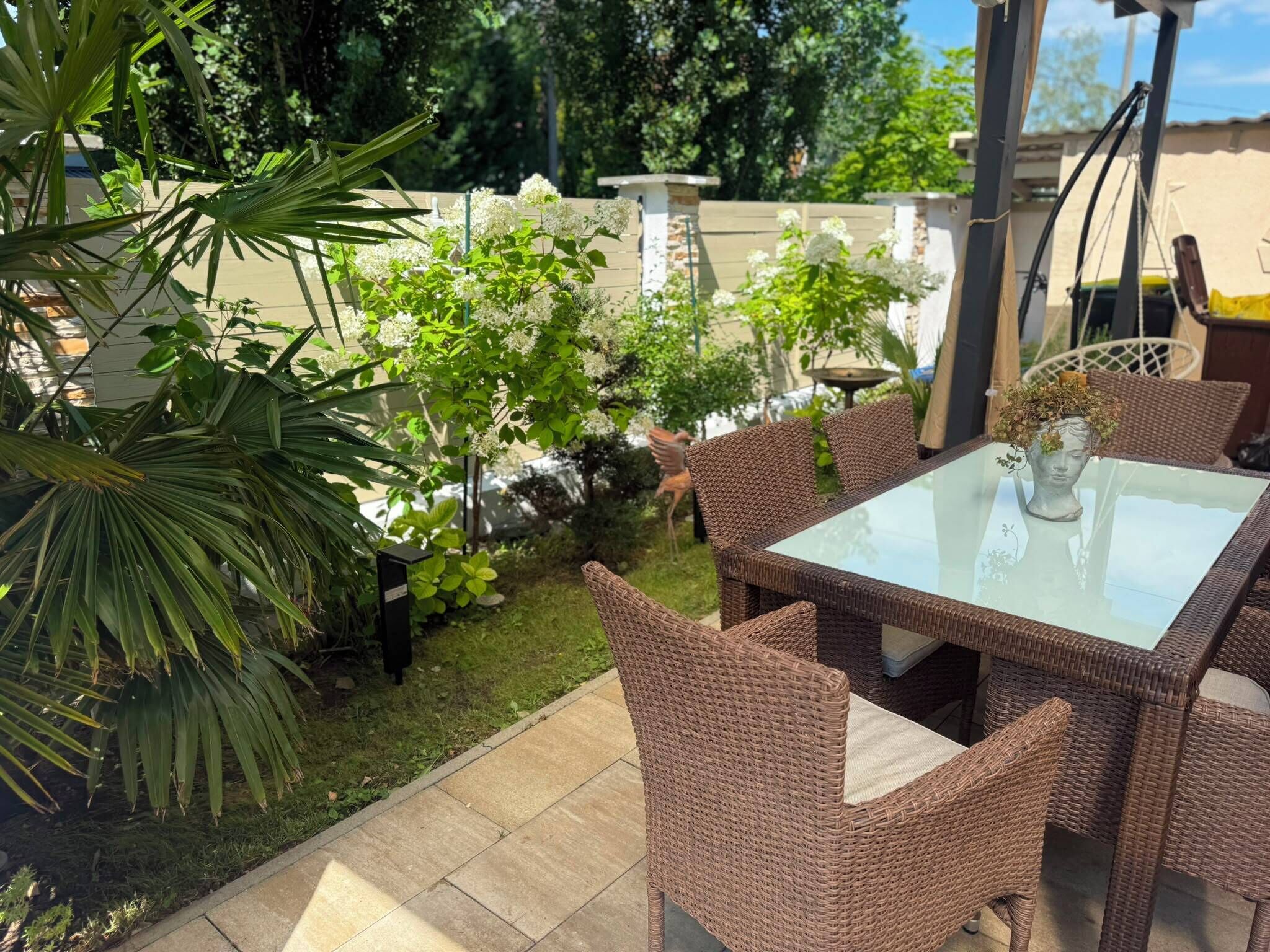 Un espace extérieur aménagé en terrasse, avec des meubles en osier, une table en verre et diverses plantes.
