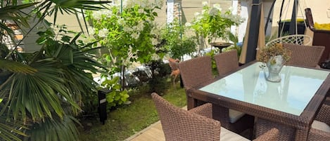 An outdoor patio area with wicker furniture, a glass table, and a variety of plants.