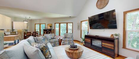 A spacious living room with a large flat-screen TV, a fireplace, and a dining area with natural light from large windows.