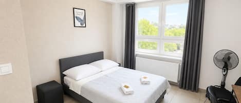 Habitación de hotel con cama, mesita de noche, una silla, ventana con cortinas y un ventilador de pared.