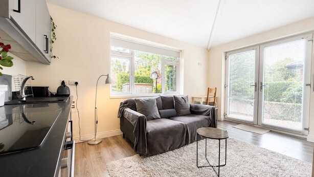 Een ruimtelijk ingerichte leefruimte met kitchenette, een grijze bank, een klein rond tafeltje en een groot raam met zonwering.