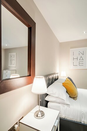 A hotel room with a bed, bedside table, lamp, and a large mirror. - Host My Nest - Modern 2 Bed Apartment by Hyde Park (London)