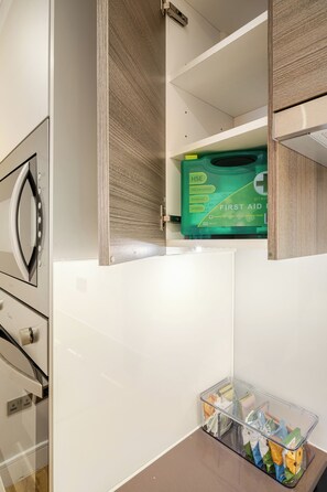 A storage compartment with a green first aid kit and a clear plastic basket with various items. - Host My Nest - Modern 2 Bed Apartment by Hyde Park (London)