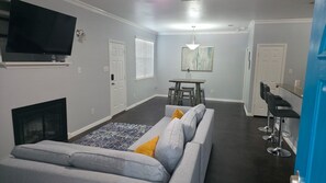 Ein Wohnzimmer mit grauem Sofa, Kamin, Wand-TV, Esstisch mit Stühlen und Küchenzeile mit Barhocker.
