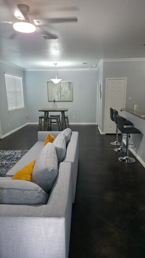 Una sala de estar moderna con un sofá gris, una mesa de comedor y una zona de cocina con taburetes de bar.