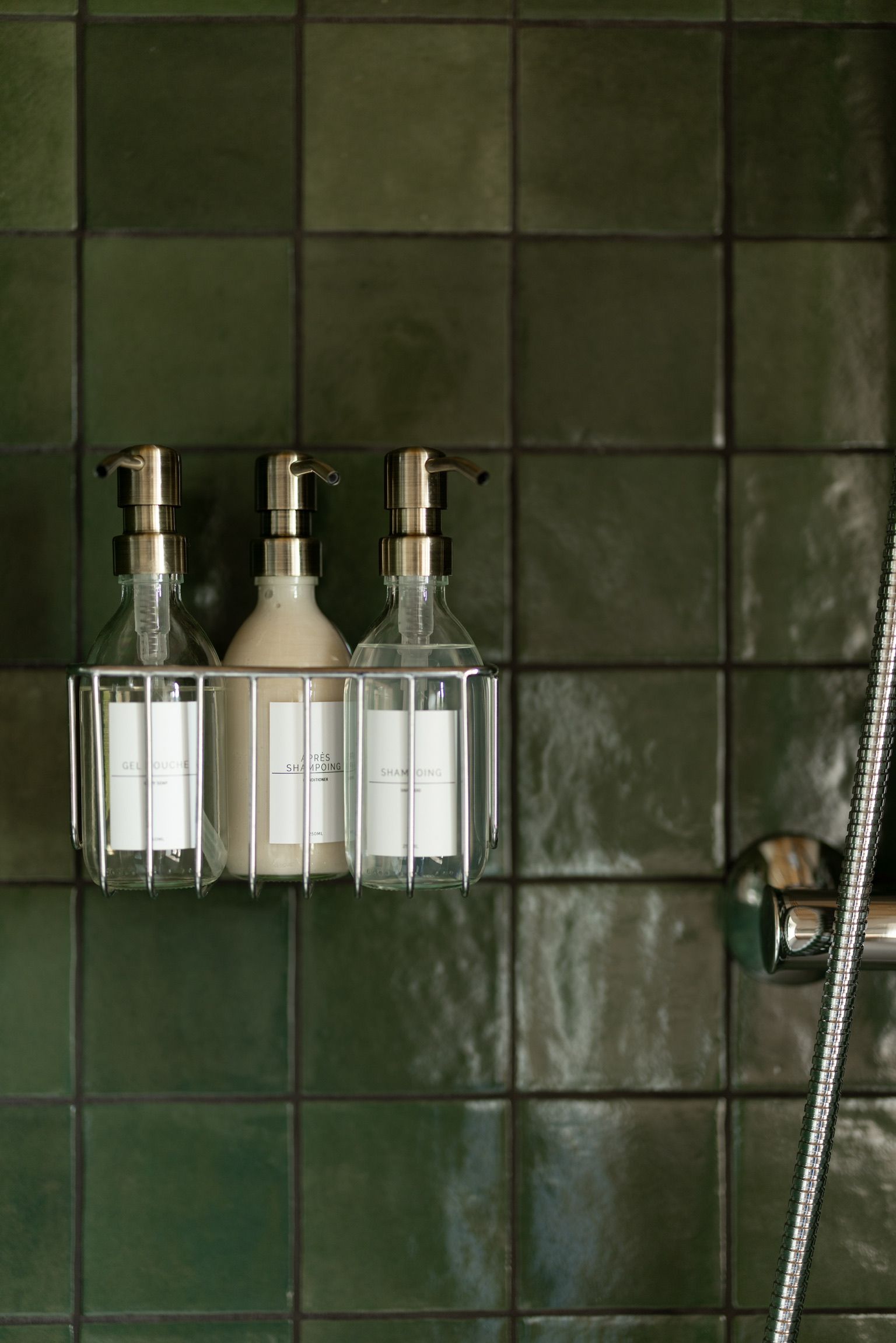 Une salle de bain avec un mur carrelé, une pomme de douche et trois bouteilles de shampoing dans un distributeur transparent.