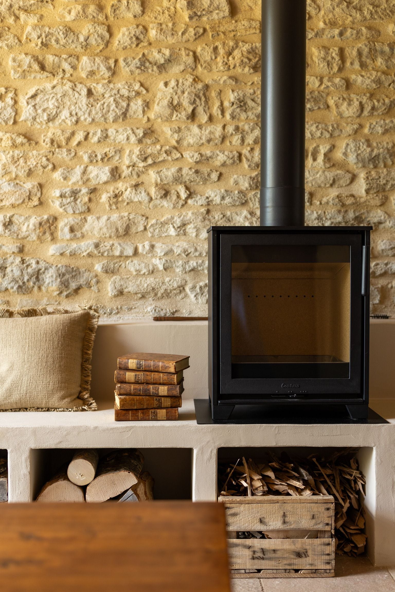 Un mur de pierres, un poêle à bois noir, une pile de livres et une caisse en bois remplie de bûches.