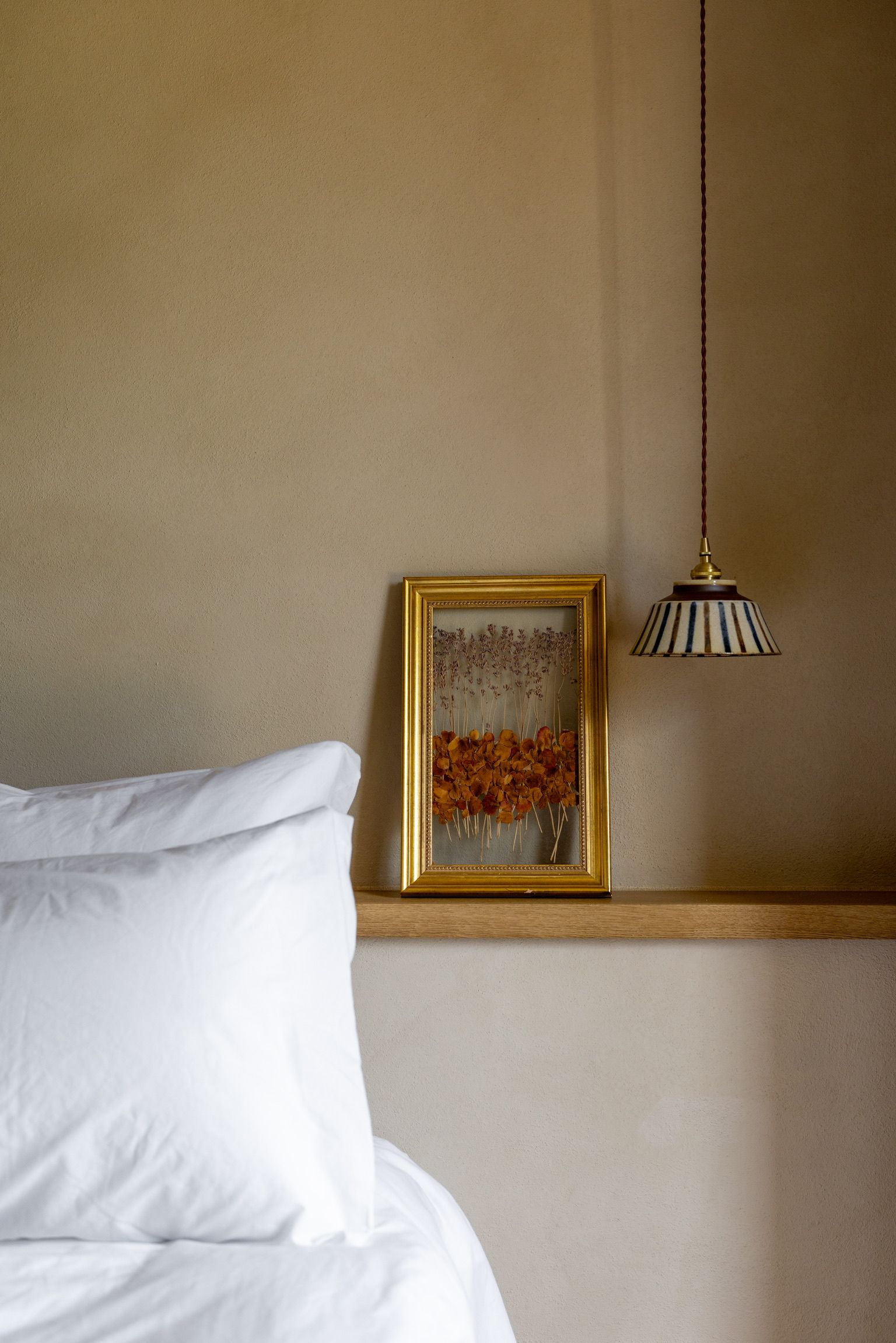 Une chambre à coucher avec un lit, un tableau encadré posé sur une étagère et une lampe suspendue.
