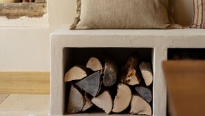 A cozy living room with a stone wall, a cushioned bench, a fireplace with logs, and a bookshelf with books and a lamp. - AUBERGE DE MARIGNA (Marigna-sur-Valouse)