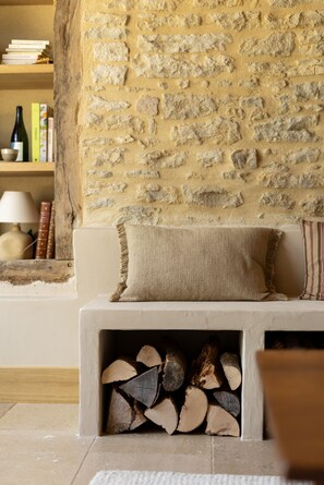 A cozy living room with a stone wall, a cushioned bench, a fireplace with logs, and a bookshelf with books and a lamp. - AUBERGE DE MARIGNA (Marigna-sur-Valouse)