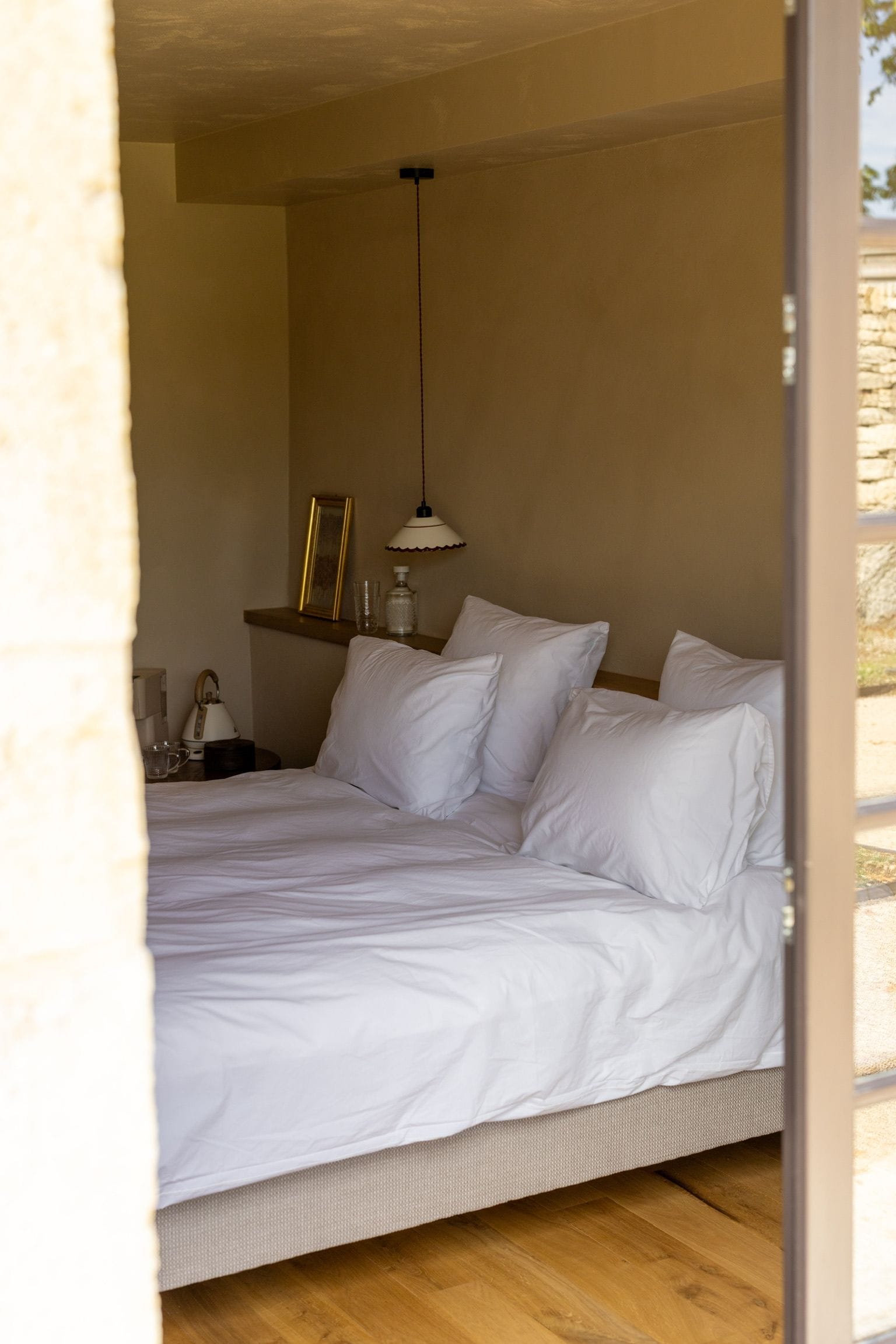 Un dormitorio con cama, mesita de noche, lámpara y una estantería de pared con objetos.