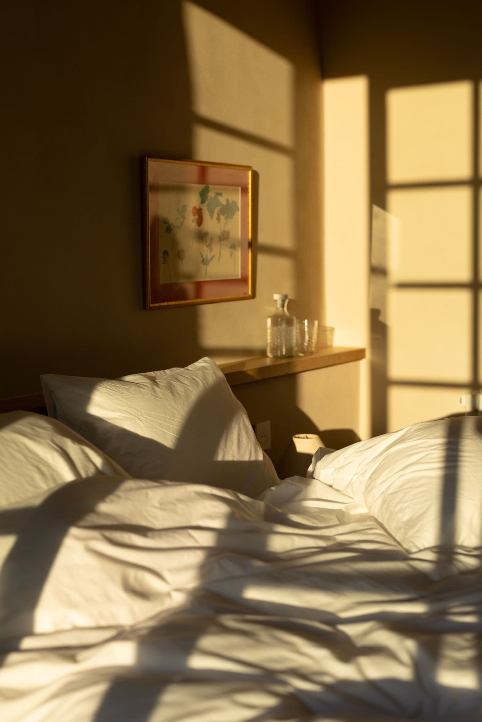 A hotel room with a bed, pillows, a bedside table with a bottle and glass, and a framed picture on the wall.