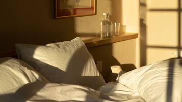 A hotel room with a bed, pillows, a bedside table with a bottle and glass, and a framed picture on the wall.