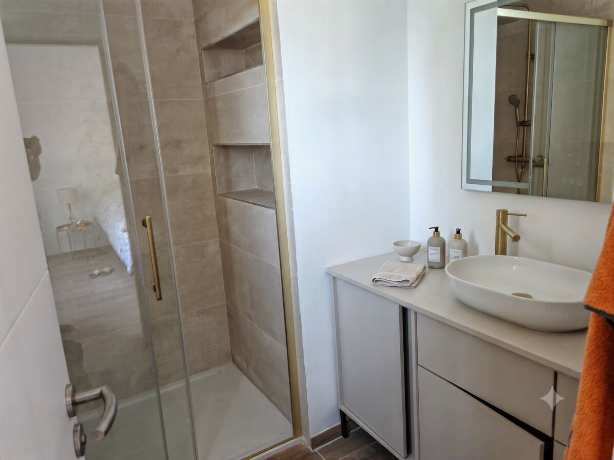 Une salle de bain moderne avec une douche à l’italienne en verre, un lavabo blanc avec un robinet doré et un miroir au-dessus du lavabo.