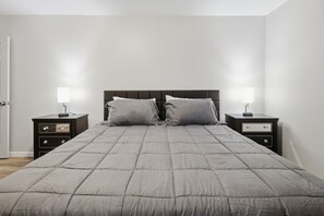 Una cama de tonos neutros con cabecera, flanqueada por dos mesitas de noche con lámparas, en una habitación sencilla con paredes blancas.