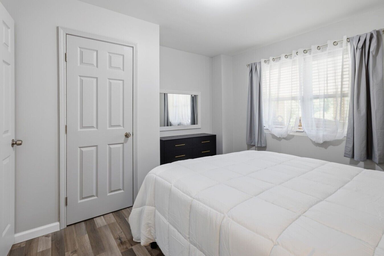 A bedroom with a bed, a dresser, a door, and a window with curtains.