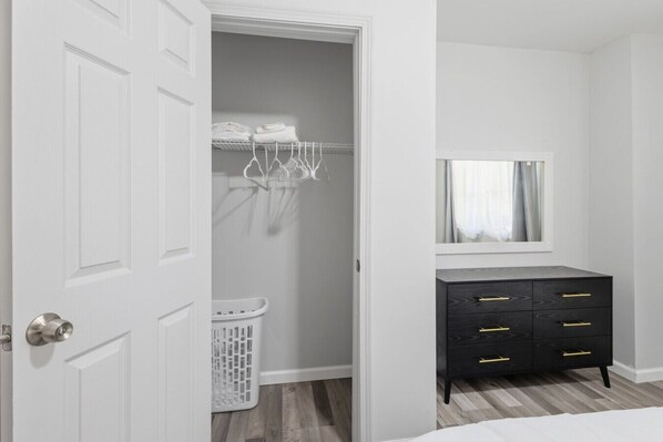 A bedroom with a white door, a black dresser with gold handles, a mirror above the dresser, and a laundry basket.