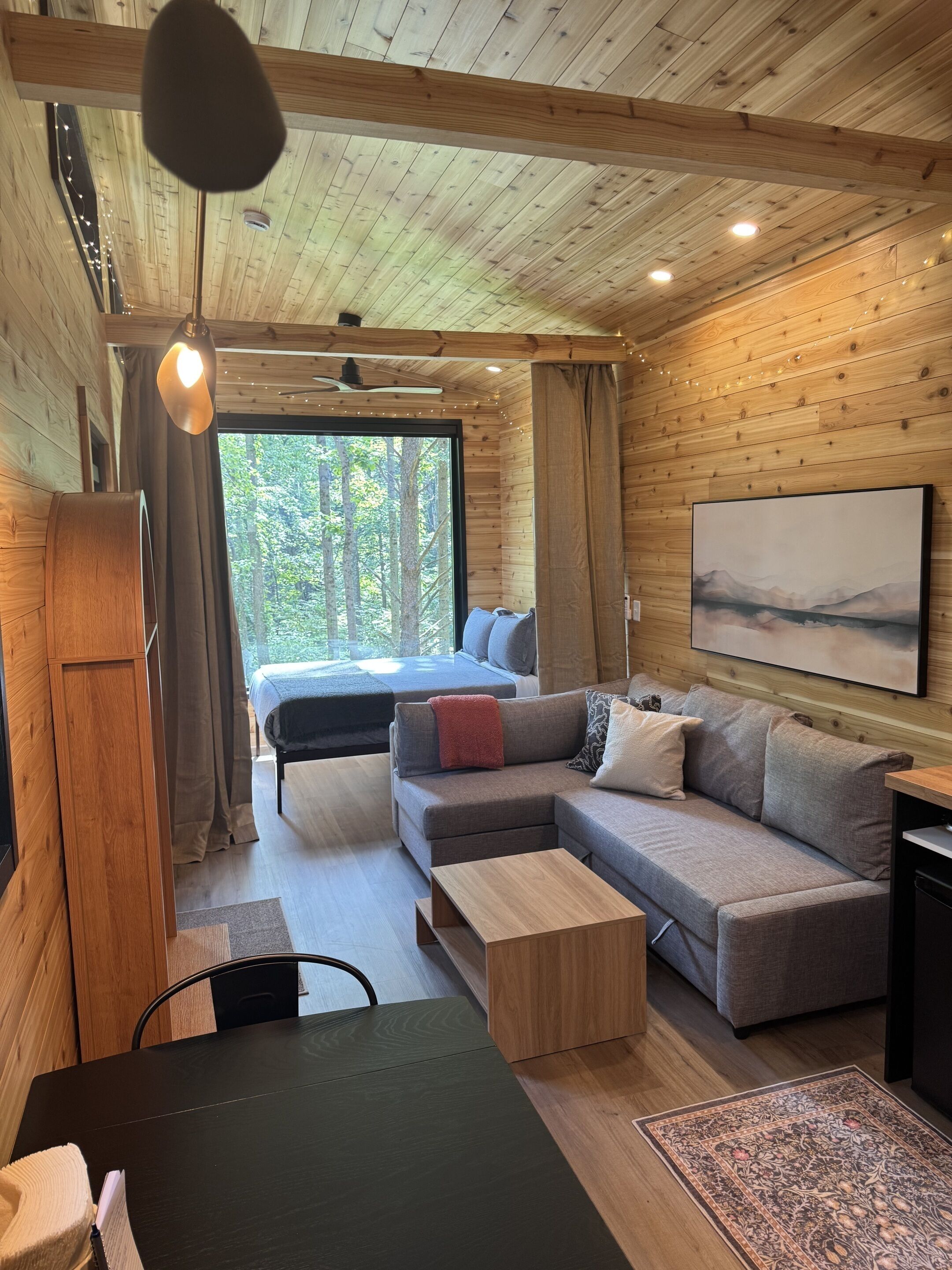 Ein gemütliches Blockhaus-Interieur mit Holzbalkendecke, einem großen Bett, einer Couch, einem Esstisch und Blick auf den Wald durch ein Fenster.