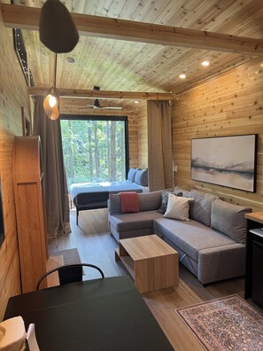 A cozy cabin interior with a wooden ceiling, a large bed, a sofa, a dining table, and a view of the forest through a window. - The Redbud Tiny House at Cyprus Trail (Rockbridge)
