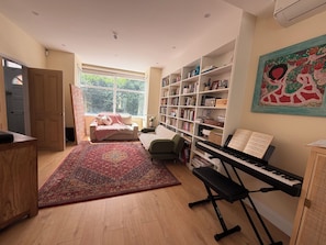 A room with a large bookshelf, a piano, a sofa, and a window with a view of trees. - West London 3-bed 3 bath family house, a/c in charming neighbourhood near centre (London)