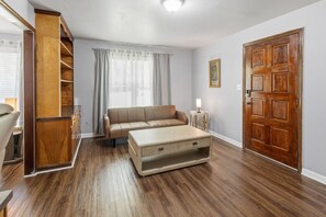 Una habitación con suelo de madera, un sofá color beige, una mesa de centro de madera, una puerta de madera y una ventana con cortinas.