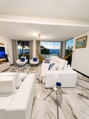 A modern living room with a white sofa set, a television, and a large window offering a view of the outdoors. - House in Mirante with Panoramic Sea View (Angra dos Reis)