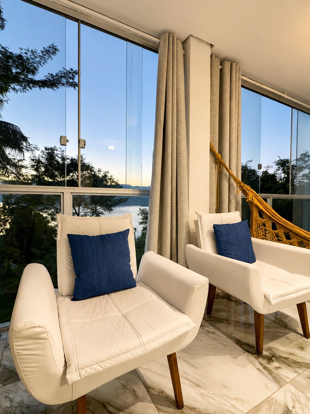 Dos sillones blancos con cojines azules, un ventanal con vistas a árboles y un cuerpo de agua, y una mecedora de madera.