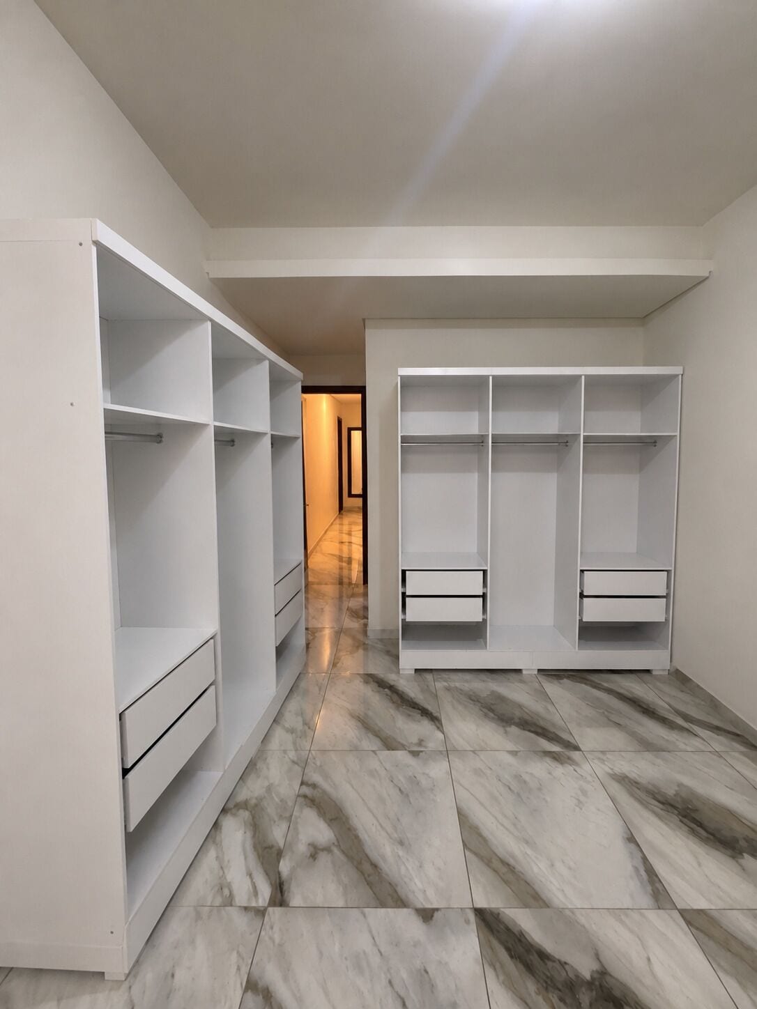 Een inloopkast met open planken en lades, een marmeren vloer, en een gang die naar een andere kamer leidt.