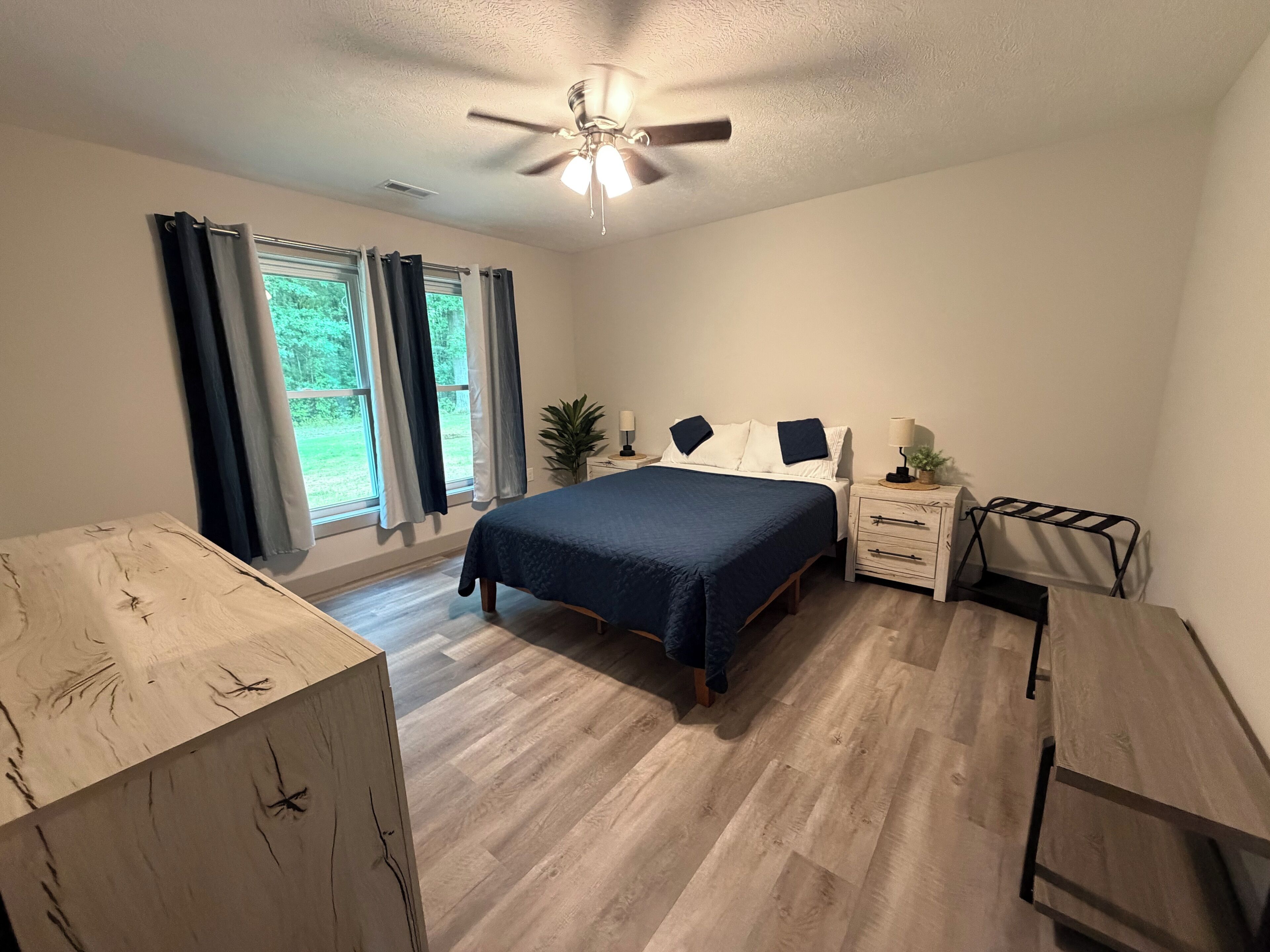 Une chambre à coucher comprenant un lit, des tables de chevet, une commode, un ventilateur de plafond et une fenêtre avec des rideaux.