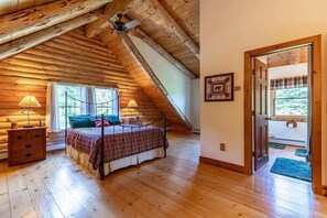 Ein Schlafzimmer mit schrägen Wänden, einem Bett mit karierter Bettdecke, zwei Nachttischlampen, einem Nachttisch und einem Fenster mit Vorhängen.
