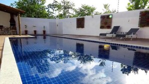 A swimming pool with tiled edges, a lounge area with chairs, and a white wall with a window. - Residencial Jardins Taperapuan (Porto Seguro)