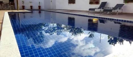 A swimming pool with tiled edges, a lounge area with chairs, and a white wall with a window.