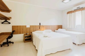 A hotel room with a desk, two beds, a chair, a small table, and a window with curtains.