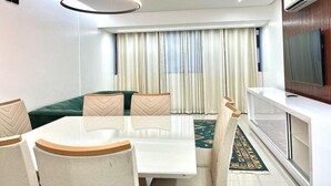 A modern living room with a dining table, chairs, a sofa, a TV, and a large window with curtains. - Residencial Jardins Taperapuan (Porto Seguro)