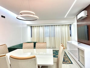 A modern living room with a dining table, chairs, a sofa, a TV, and a large window with curtains.