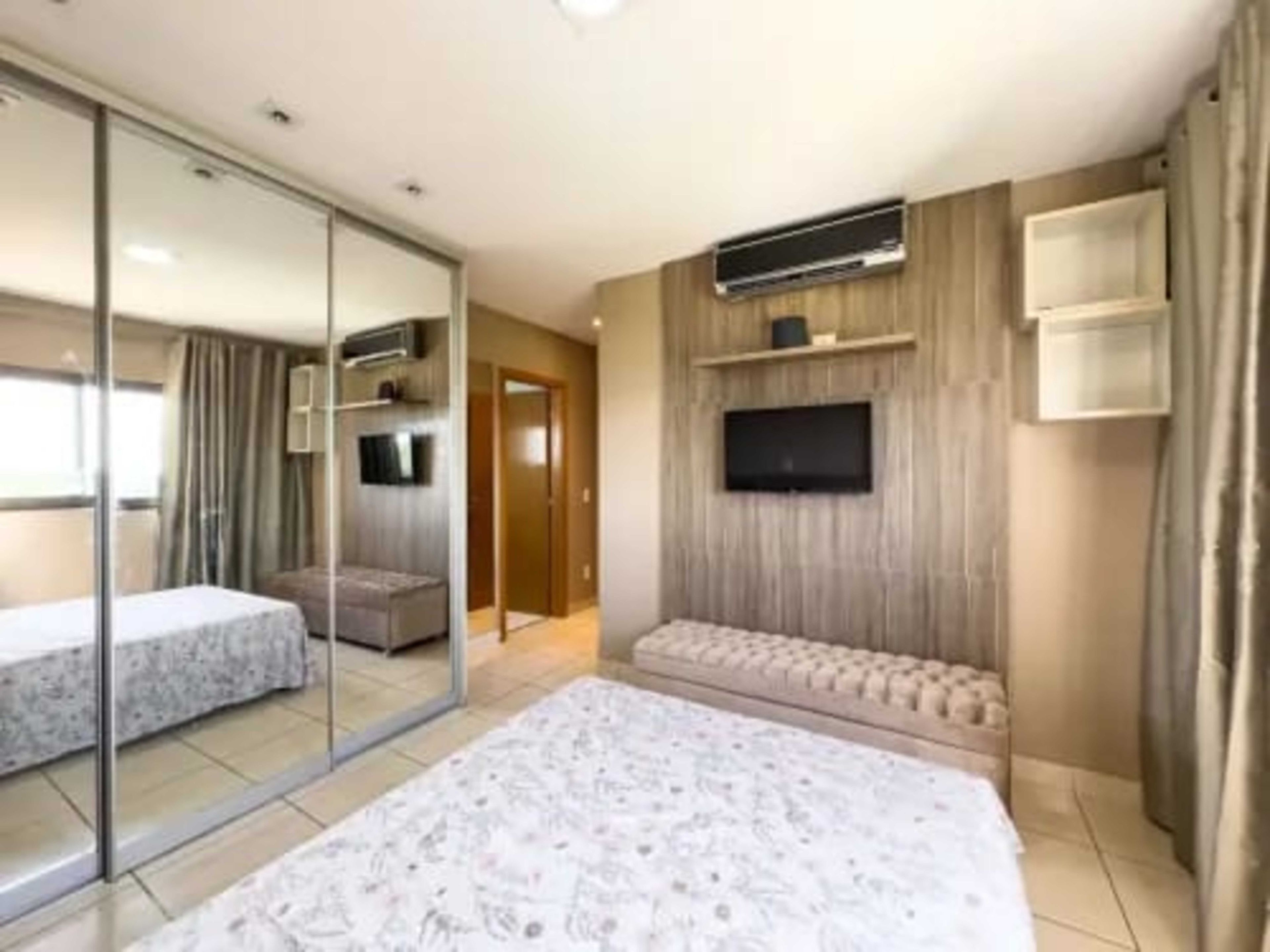 A modern hotel room with a large bed, a sofa, a TV mounted on the wall, and a sliding glass door leading to a balcony.