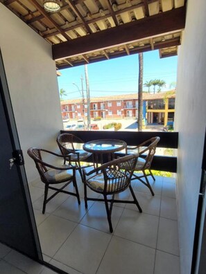 Uma varanda com cadeiras de vime e uma mesa redonda pequena, com vista para uma rua com edifícios e palmeiras.