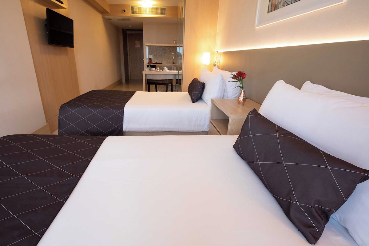 A hotel room with two beds, a small desk, a mirror, and a kitchenette in the background.