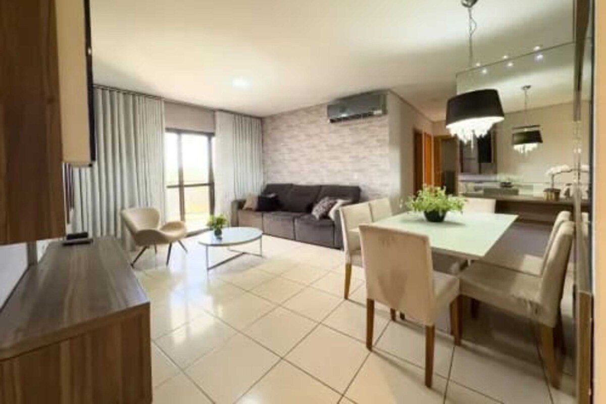Uma sala de estar moderna com sofá, mesa de jantar com cadeiras e área de cozinha com luminária suspensa.