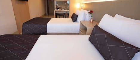 A hotel room with two beds, a small table, a chair, a TV mounted on the wall, and a kitchenette in the background.