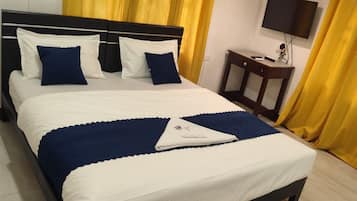 Cama bem arrumada com roupa de cama branca e azul, mesa de cabeceira com abajur e cortinas amarelas.
