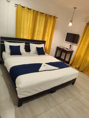 A neatly made bed with white and blue bedding, a bedside table with a lamp, and yellow curtains. - Hotel Aidan Residency Edappally (Kanayannur)