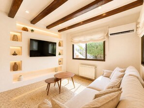 A modern living room with a flat-screen TV, built-in shelves with decorative items, a wooden ceiling, a radiator, and a large window with a view of greenery. - Villa Las Marinas CF with Private Pool near the Beach in Dénia (Denia)