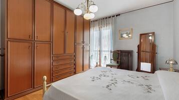Un dormitorio con un armario grande, una cama con colcha estampada, una mesita de noche con lámpara y una ventana con cortinas de gasa.
