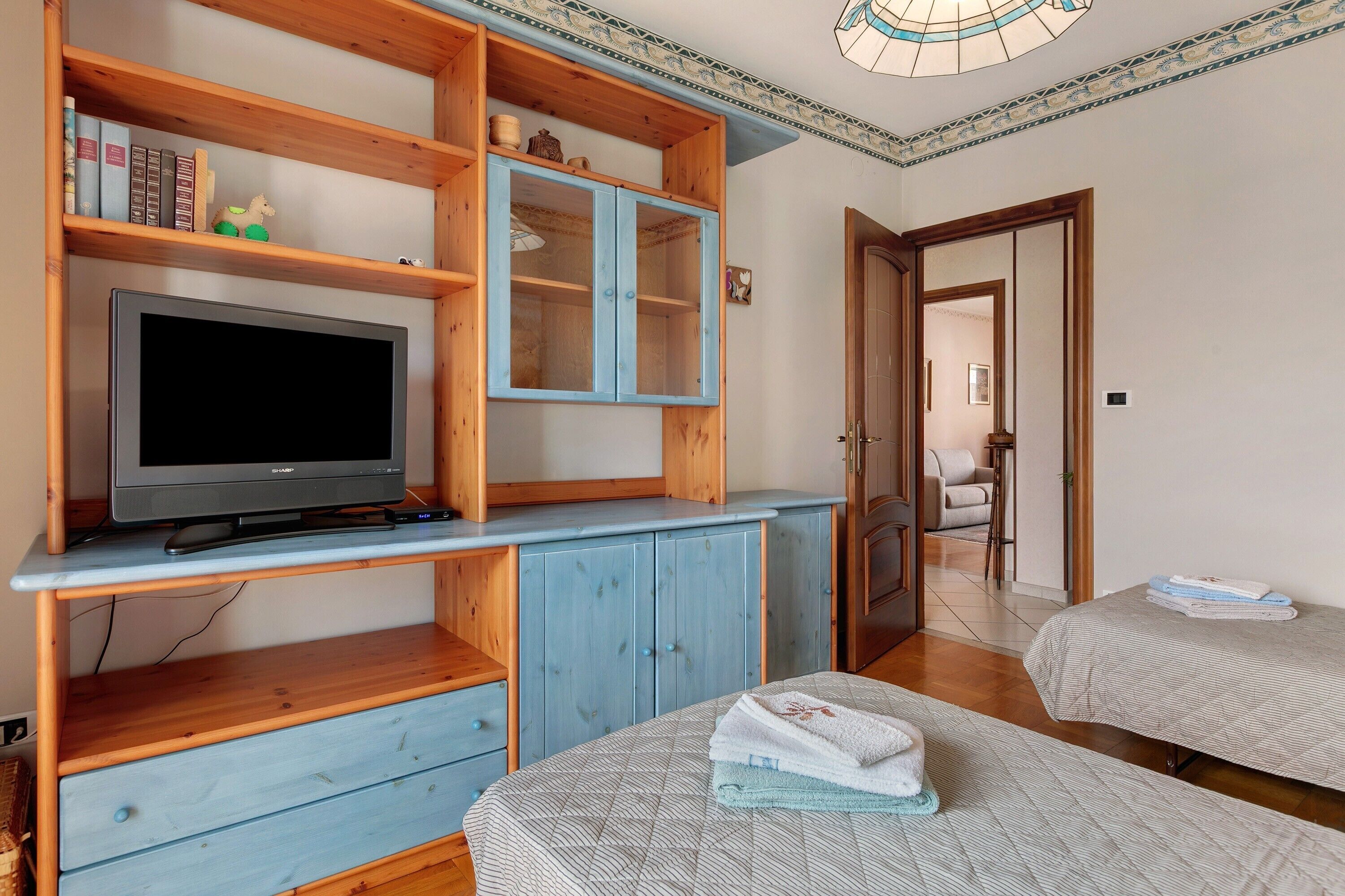 Une pièce avec un téléviseur posé sur un meuble en bois, un buffet bleu aux portes vitrées et deux lits avec des serviettes pliées.