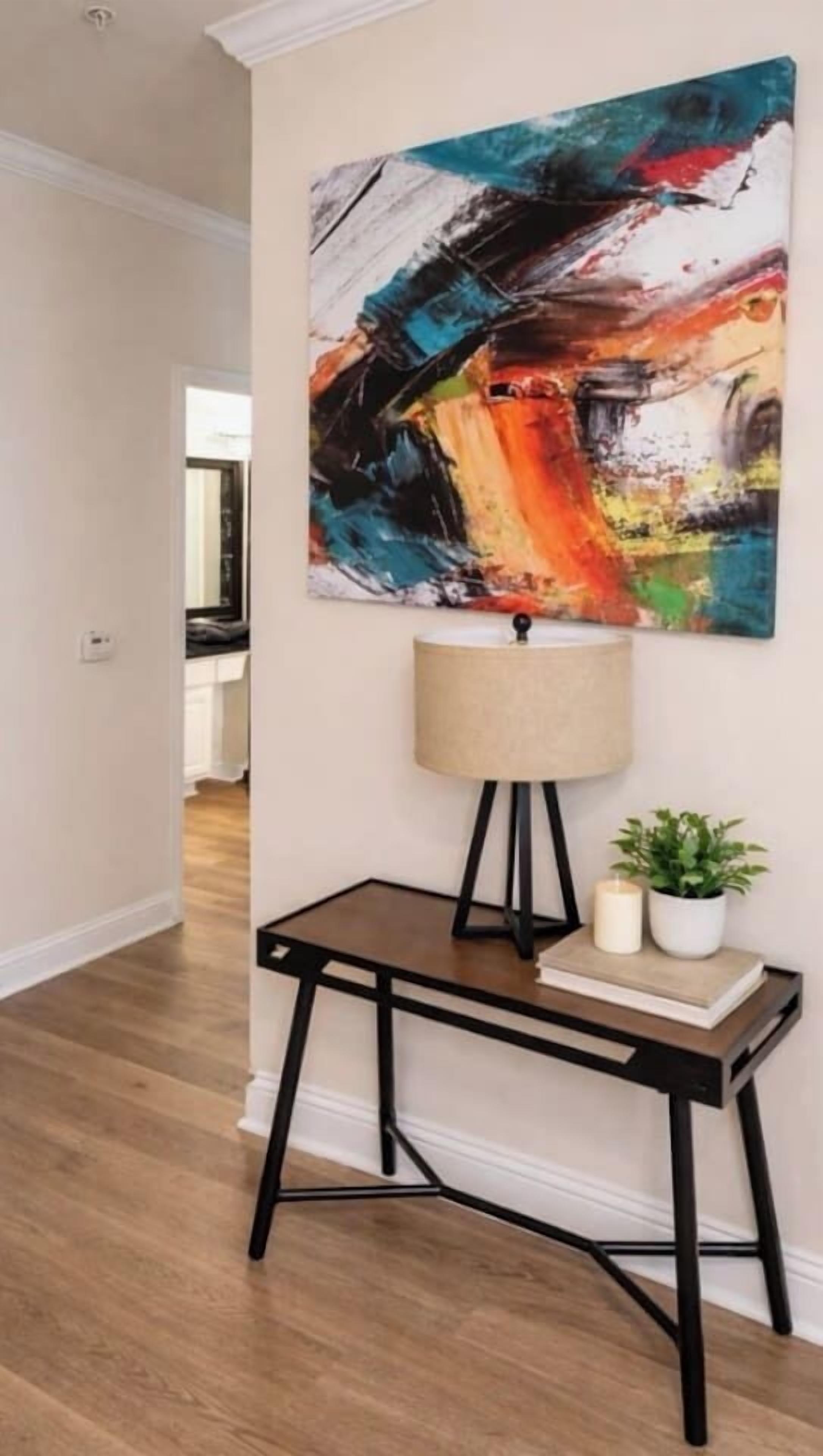 Ein Zimmer mit Holzboden, einem Schreibtisch mit Lampe, einer Topfpflanze und einem farbenfrohen, abstrakten Gemälde an der Wand.